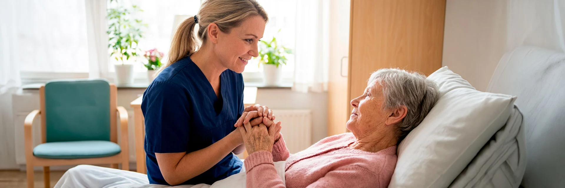 Undersköterska i blå arbetskläder sitter vid sängkanten och håller en äldre kvinnas händer. De ler mot varandra i ett ljust vårdrum med fönster och gröna växter i bakgrunden.